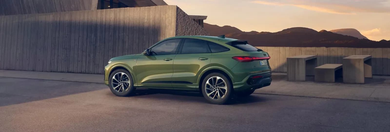 Audi SUV coupé verde in sosta al tramonto davanti a un muro in cemento, con montagne sullo sfondo
